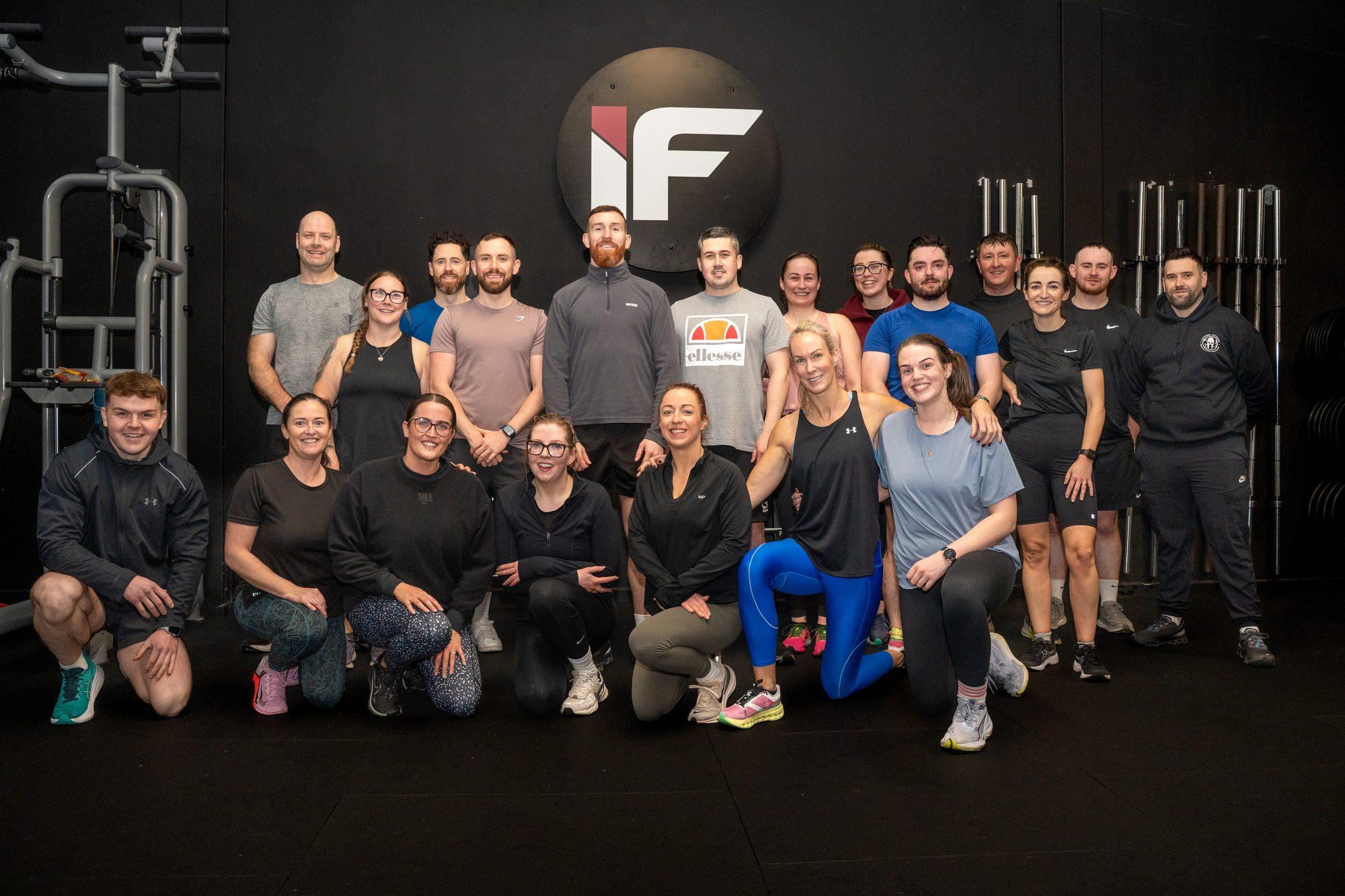 A group of Intensity Fitness members and coaches smiling together on the gym floor, beneath the IF logo on the wall