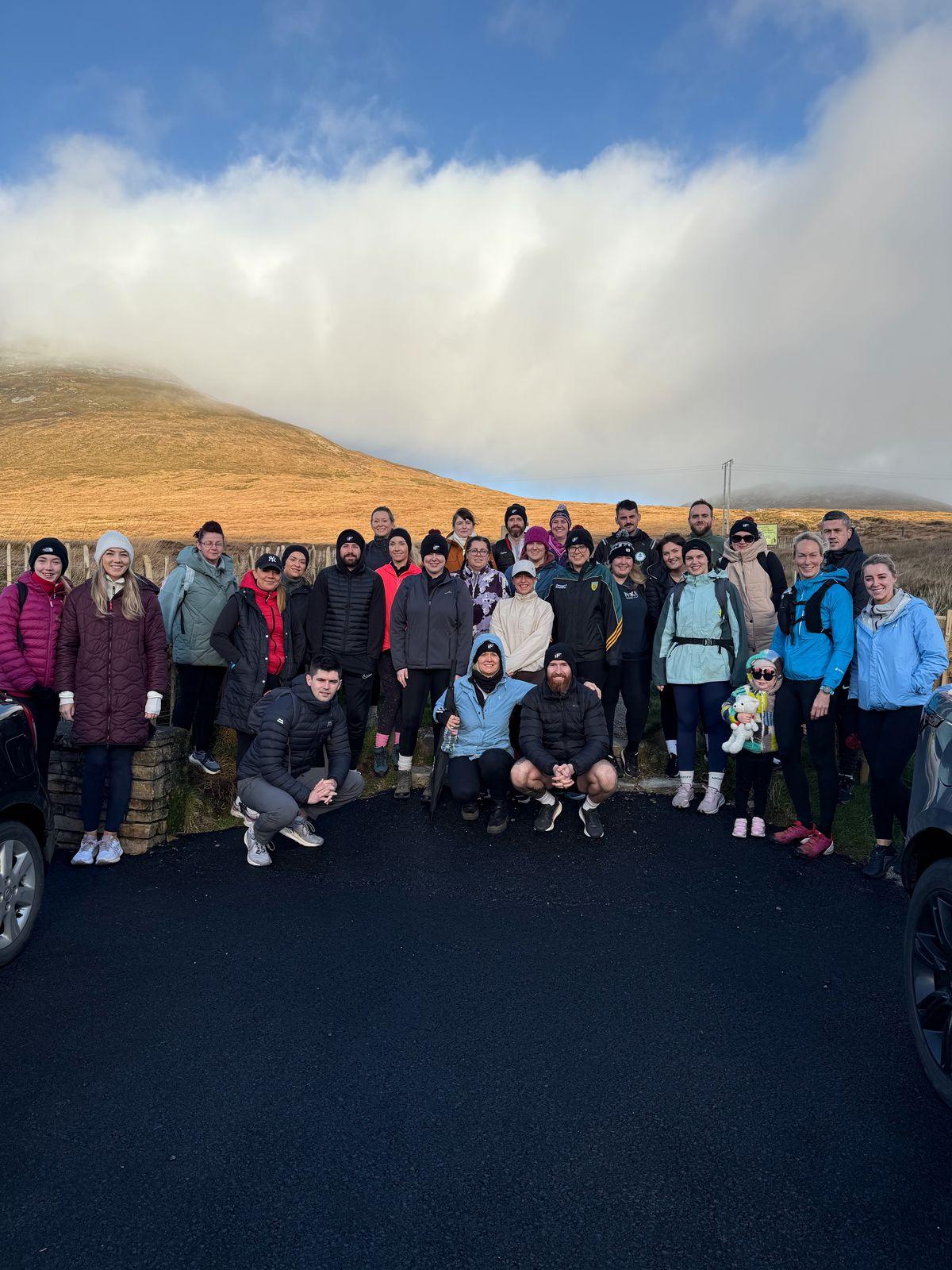 A large group of Intensity Fitness members and coaches, adults and one young child, posing together on a hillside road with a mountain behind them